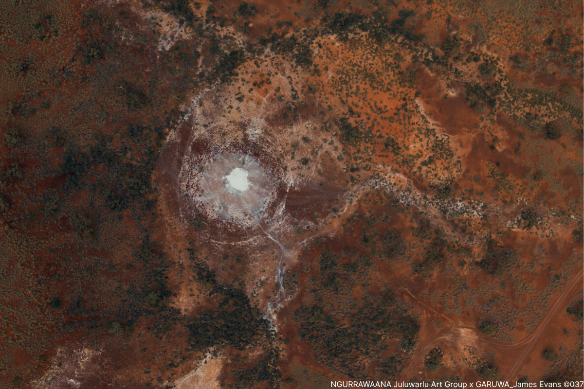 Aerial view of red desert landscape with a white circular salt formation at the centre, surrounded by scattered vegetation.